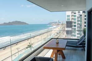 Varanda com mesa de madeira e duas cadeiras de descanso com vista para uma praia e oceano.