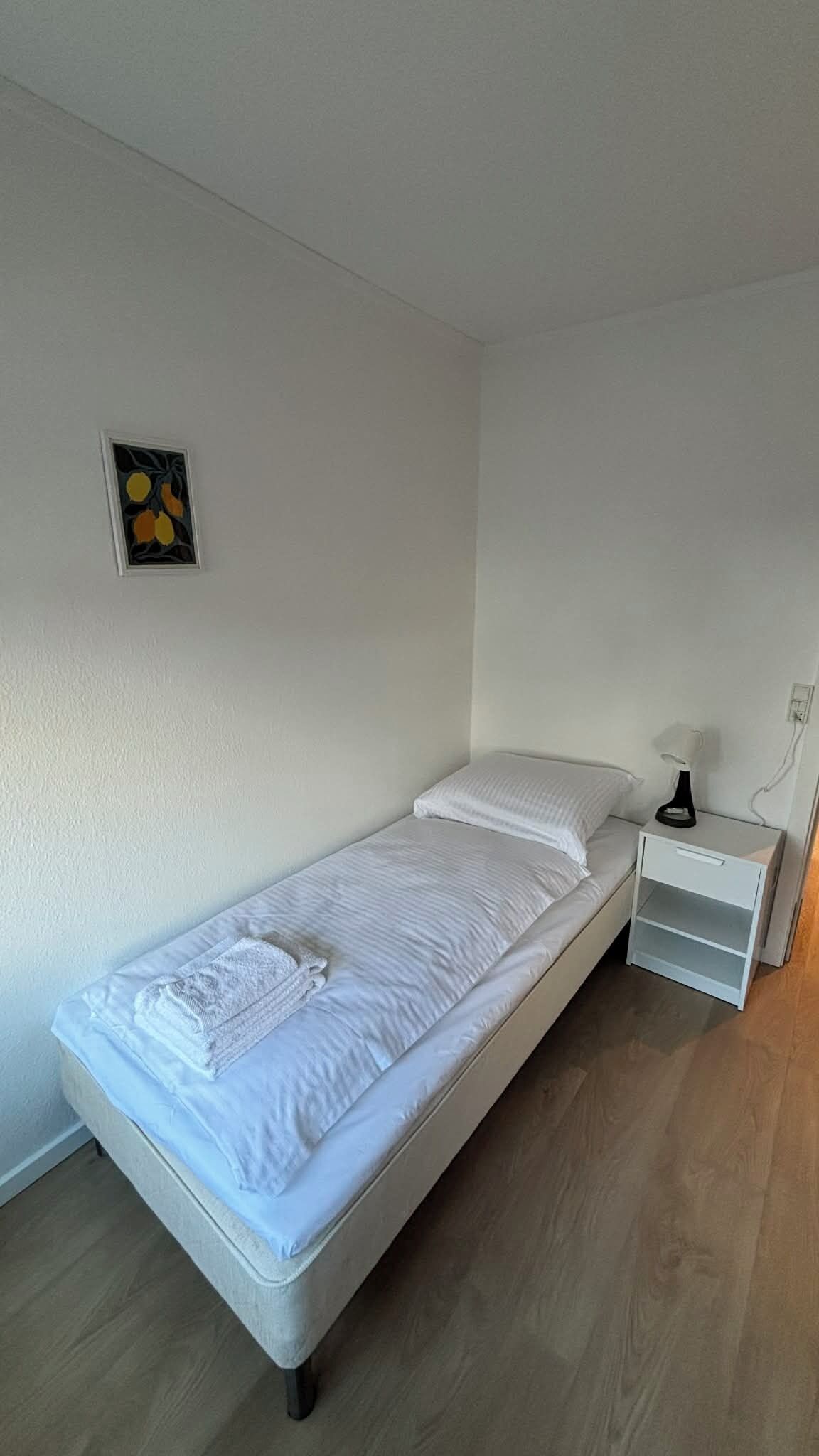 Een eenpersoonsbed met witte lakens en kussens, een nachtkastje met een lamp, en een klein ingelijst foto aan de muur.