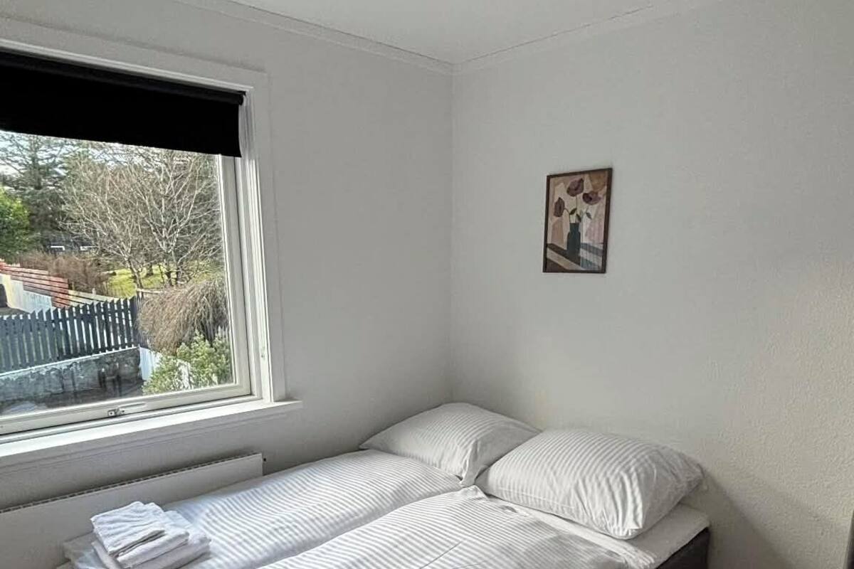 Un petit lit bien fait, avec du linge de lit blanc et des oreillers, dans une pièce aux murs blancs, dotée d’une fenêtre et d’un radiateur.