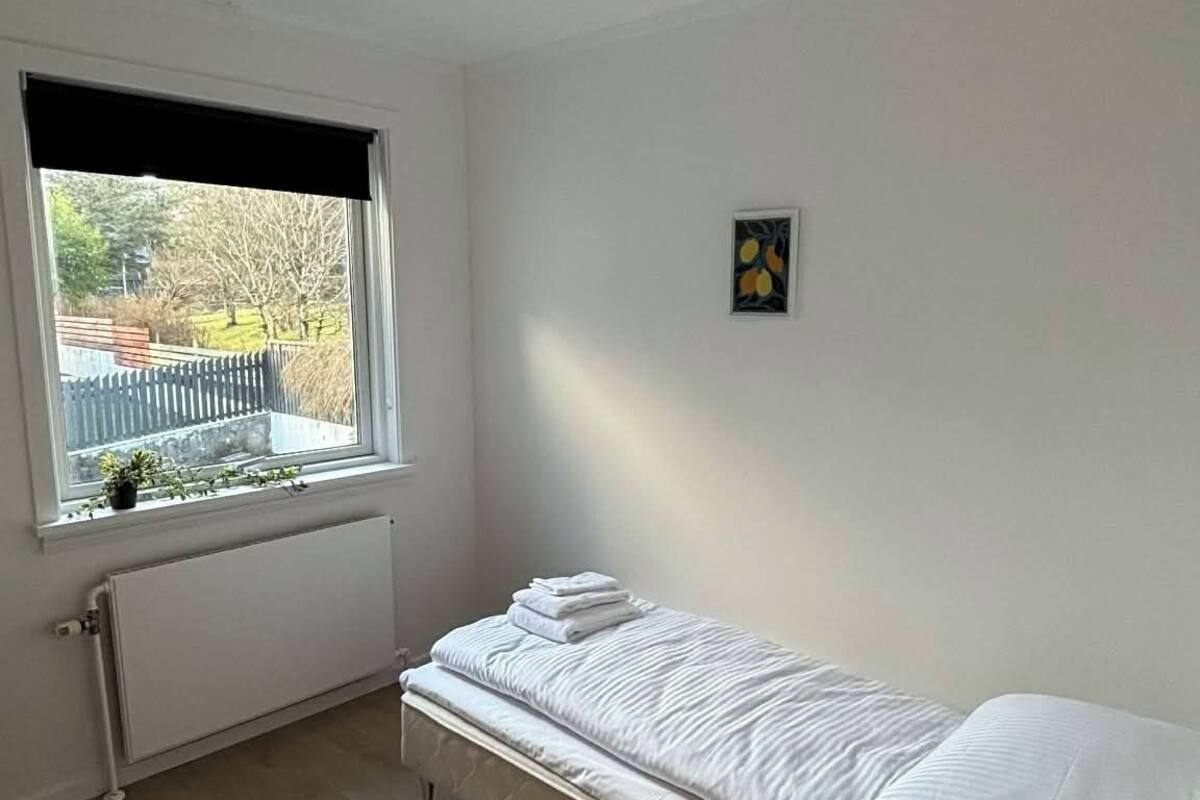 Une petite chambre à coucher au style minimaliste, avec un lit simple, un radiateur, une fenêtre donnant sur l’extérieur et un petit tableau décoratif au mur.