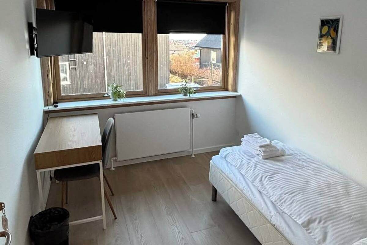 Une petite pièce bien éclairée, avec un lit simple, un bureau et une chaise, un radiateur et une fenêtre donnant sur une clôture et des arbres.