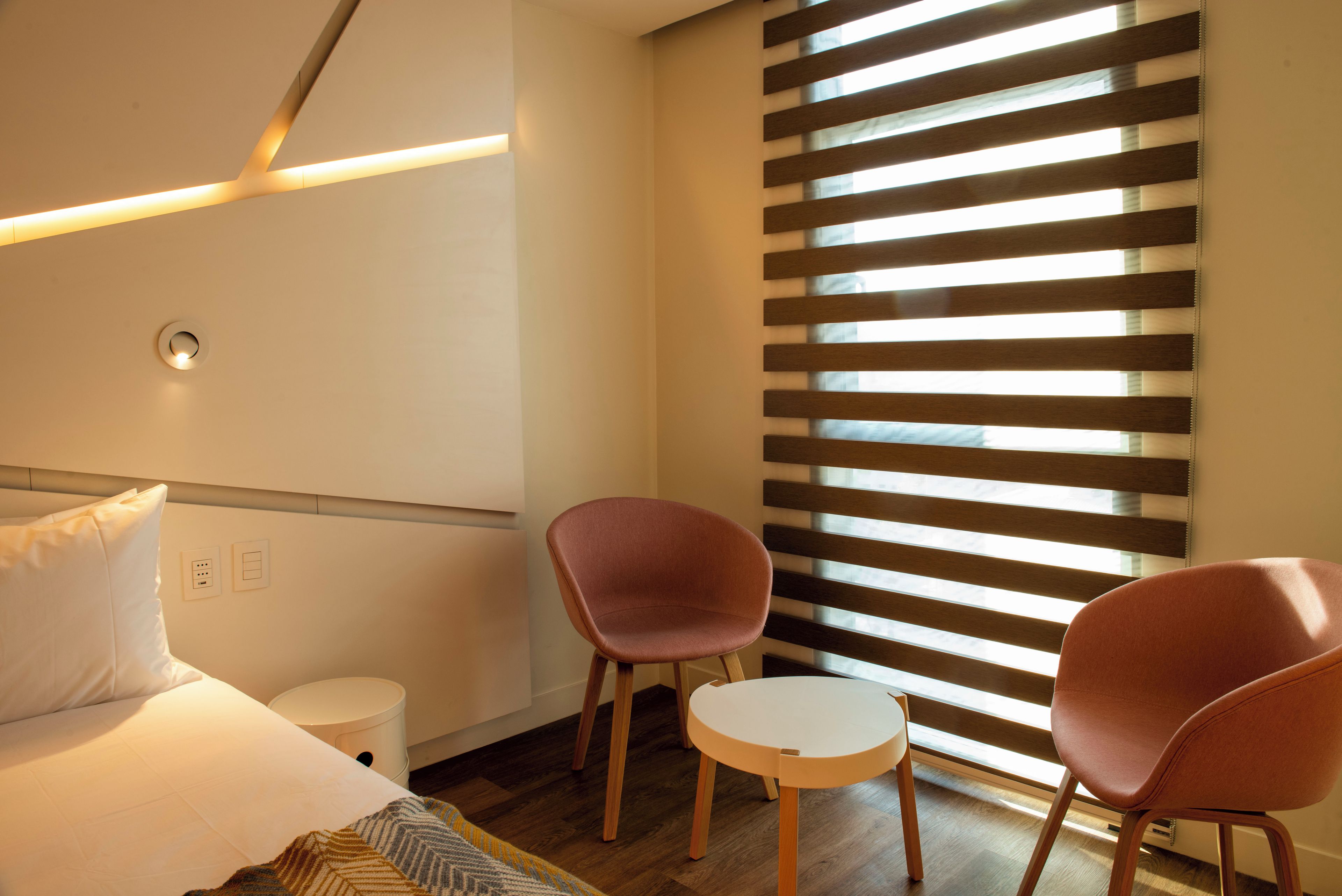 A hotel room with a bed, two chairs, a small table, a bedside table, a waste bin, and a window with blinds.