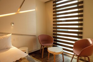 A hotel room with a bed, two chairs, a small table, a bedside table, a waste bin, and a window with blinds. - Hotel Nodo (Santiago)