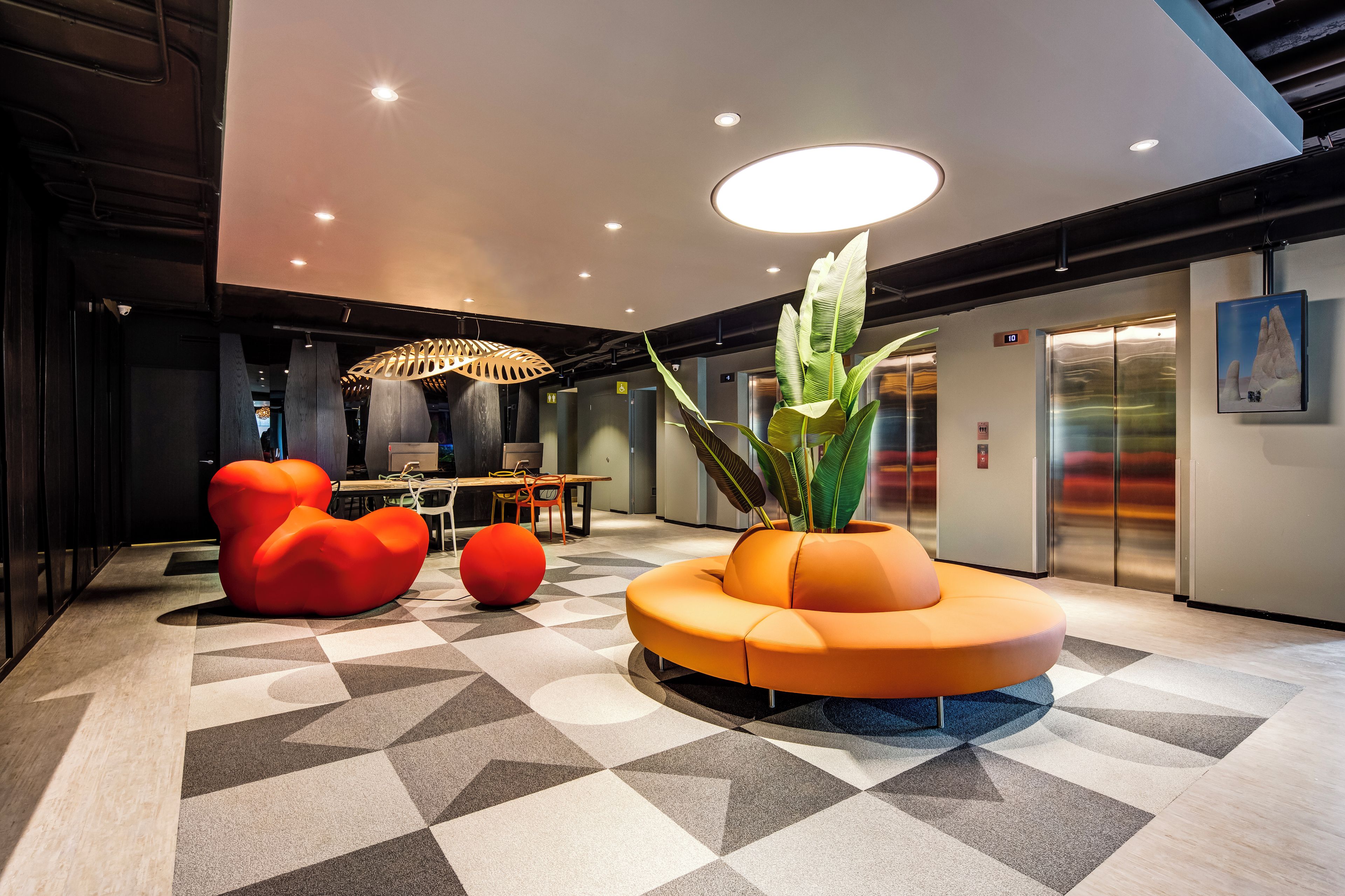 A modern lobby with a geometric patterned floor, a central orange seating area, and an elevator.