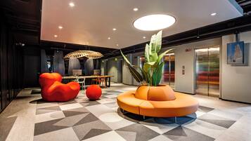 A modern lobby with a geometric patterned floor, a central orange seating area, and an elevator.