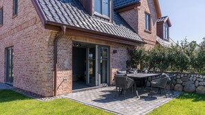 Een bakstenen huis met een schuin dak, een terras met buitenmeubilair en een stenen muur.