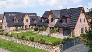 Een rij van drie bakstenen huizen met schuine daken, gelabeld 'Dorfhüs 1', 'Dorfhüs 2' en 'Dorfhüs 3'. Elk huis heeft een kleine garage en een voortuin met een stenen rand.
