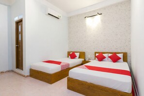 Une chambre d’hôtel avec deux lits en bois, du linge de lit blanc, des touches de couleur rouge, un climatiseur fixé au mur et un papier peint à motifs.