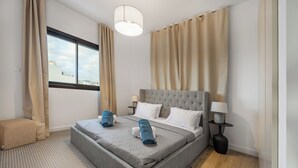 Una habitación de hotel moderna con una cama grande, cortinas color beige, un ventanal con vista a la ciudad, una lámpara redonda y una mesita de noche pequeña con lámpara.