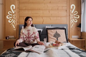 Uma pessoa sentada na cama com um livro, uma bandeja com doces e uma xícara.