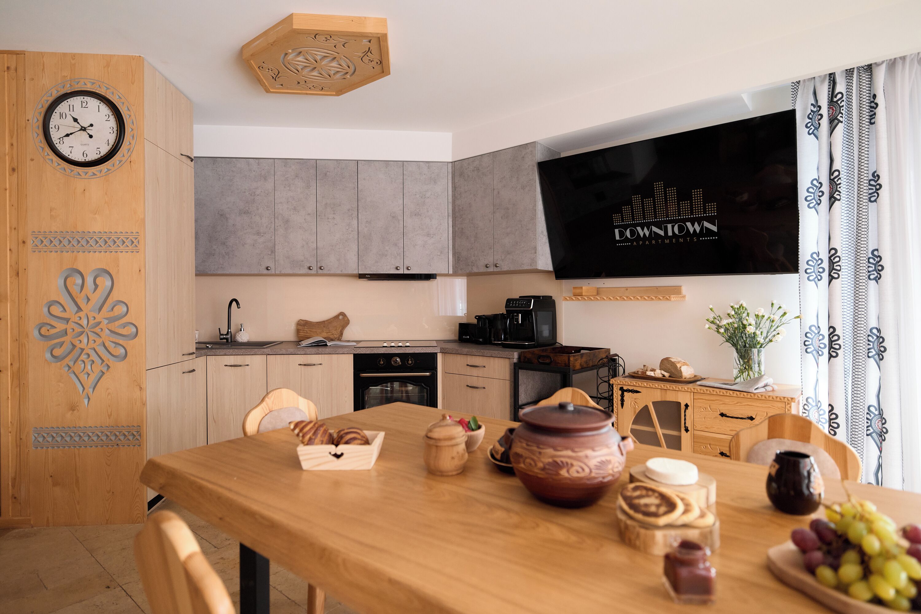 A modern kitchen with grey cabinets, a wooden dining table with bread, a large TV, and a clock on the wall.