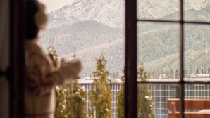 Una persona de pie junto a una ventana con vistas a montañas nevadas y un lago.