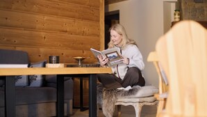 Una persona sentada en una silla con una manta de piel, leyendo un libro en una habitación con paredes de madera y un sofá.