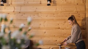 Interior de una cabaña de madera con una mujer cortando una tarta en una mesa de madera.