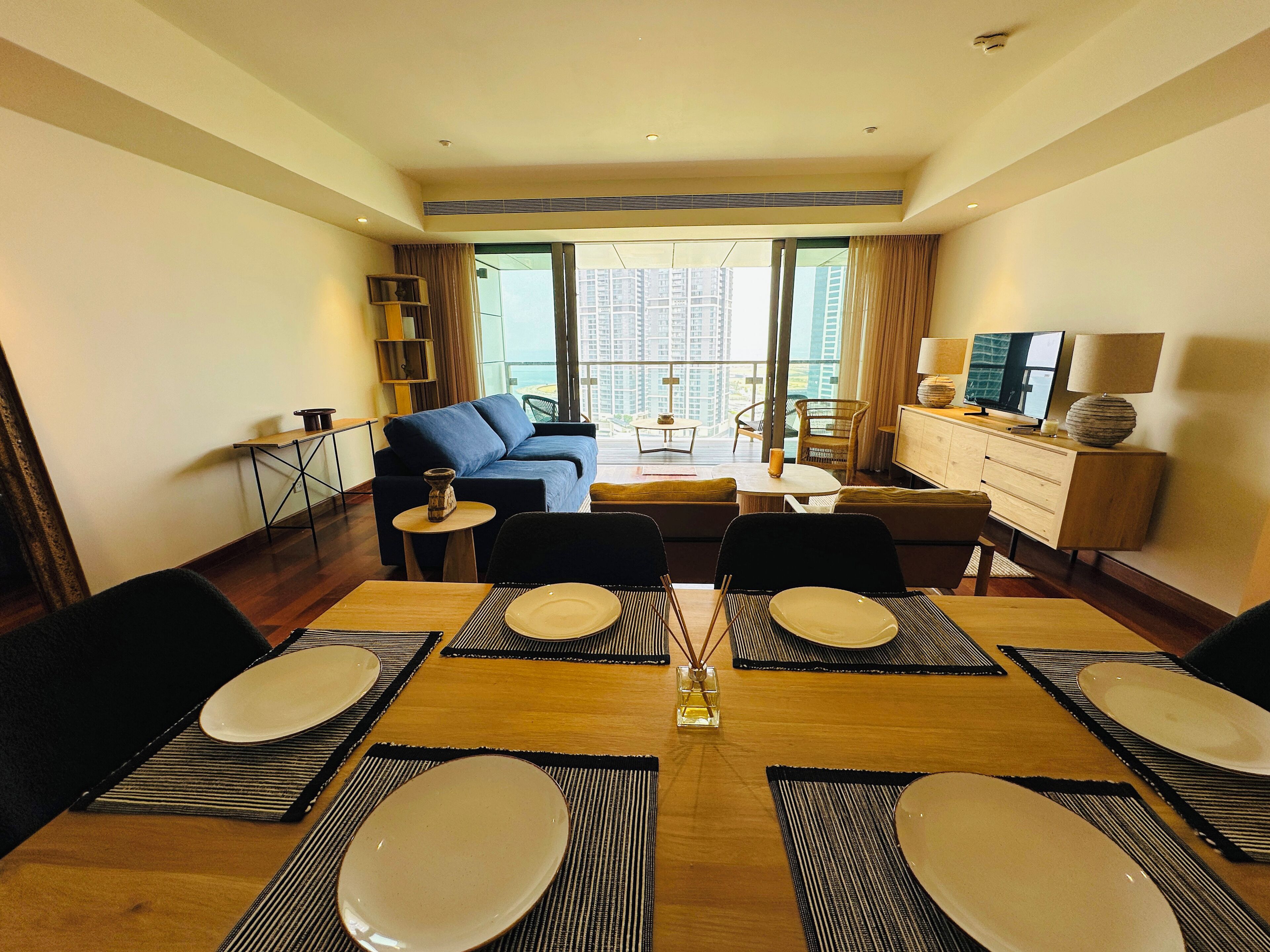 A modern hotel room with a dining area, a sofa, a TV, and a balcony with a city view.