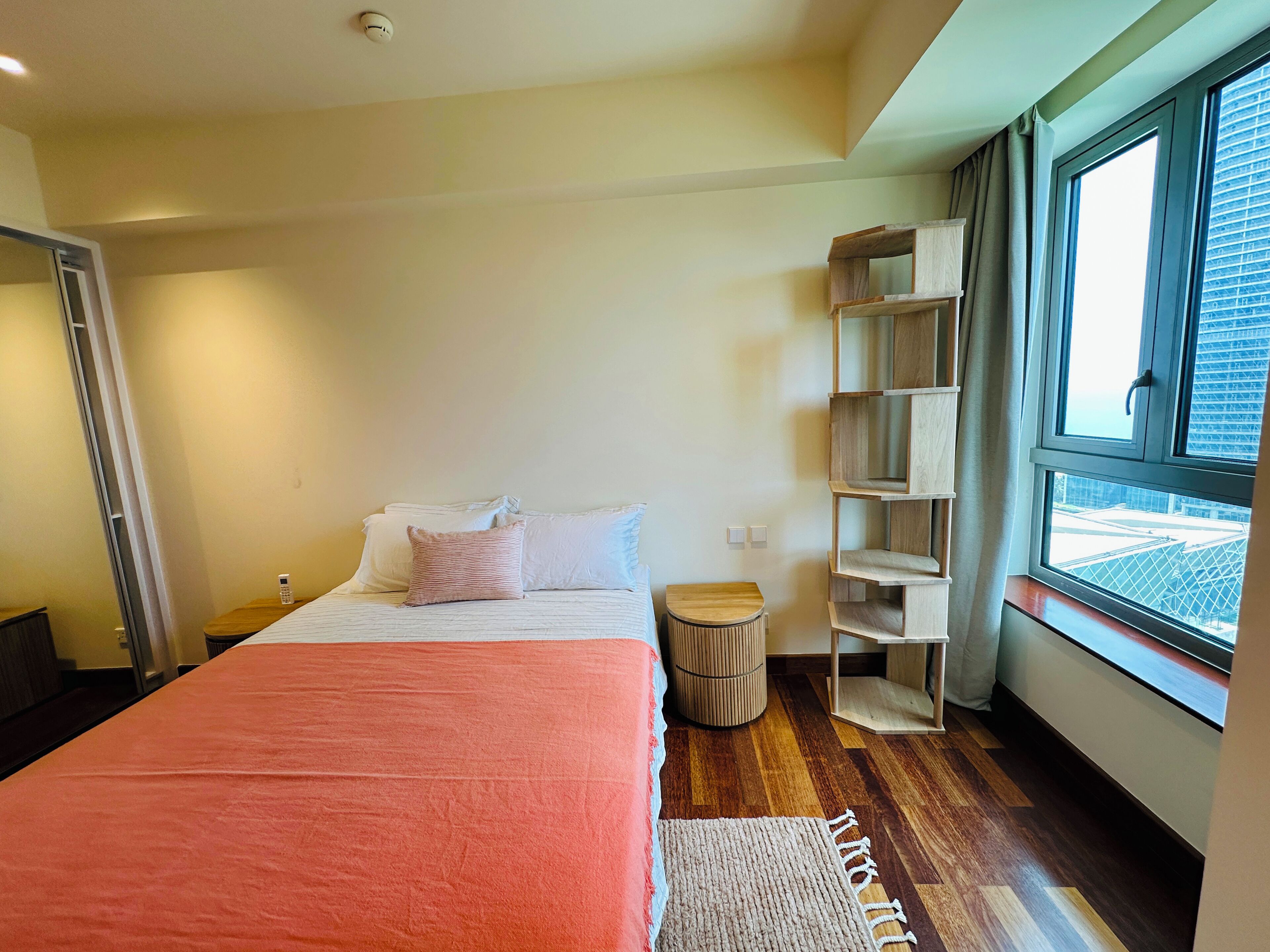 A bedroom with a bed, bedside tables, a small round stool, a wooden shelf unit, and a window with a view of buildings.