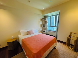 A bedroom with a bed, a bedside table, a small round table, a window with a view of buildings, and a wooden ladder.