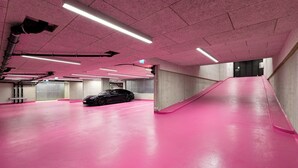 Un parking au sol recouvert d’un revêtement et aux murs de couleur rose, avec une voiture noire et des piliers en béton.