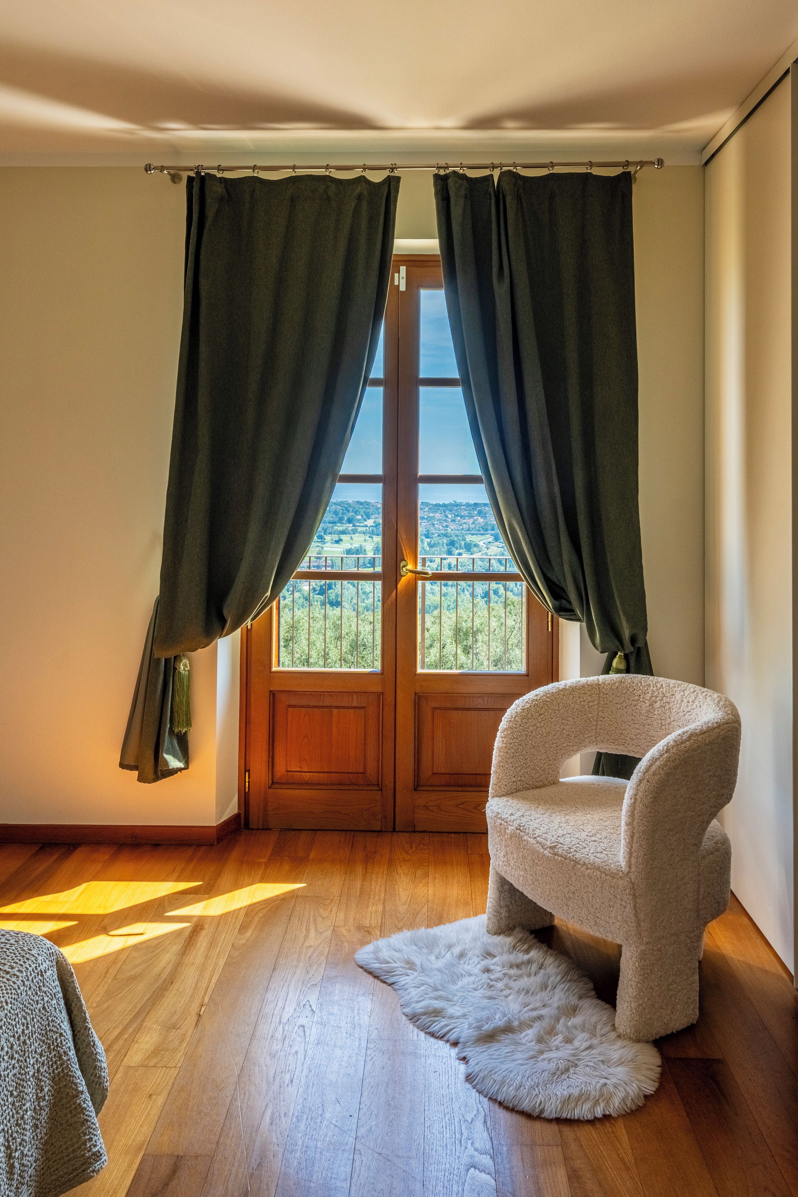 Une pièce avec un plancher en bois, une chaise blanche, une vue à travers une porte avec des rideaux et une fenêtre donnant sur un paysage verdoyant.