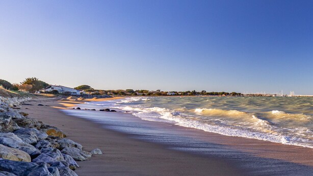 Una playa con olas, costa rocosa y edificios a lo lejos.