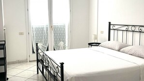 A bedroom with a bed, a nightstand, a lamp, a window with curtains, and a view of buildings outside. - P081 - Porto Recanati, new three-room apartment with balcony (Porto Recanati)