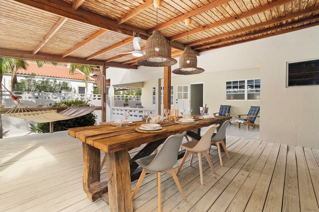 A wooden dining table set with chairs, a hammock, and a view of a house with a red roof.