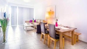 Ein modernes Wohnzimmer mit einer Couch, Couchtisch, Esstisch und Stühlen. An der Wand hängt ein Gemälde und auf dem Tisch steht eine Vase mit Pflanzen.