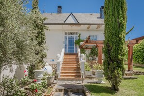 A house with a staircase leading to a blue door, surrounded by greenery and potted plants.