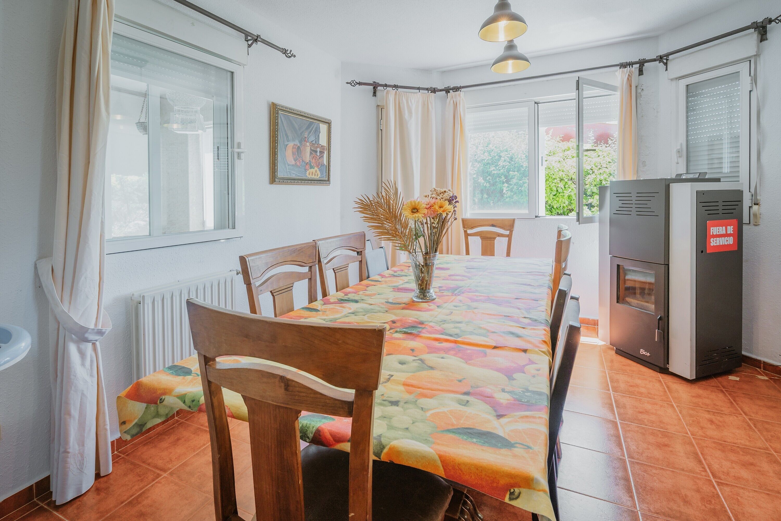 A dining area with a table, chairs, a vase with flowers, a radiator, a window, and a kitchen area with a refrigerator.