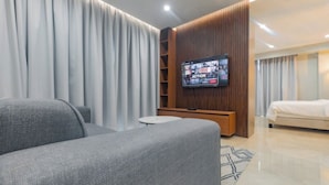 A modern hotel room with a large flat-screen TV mounted on a wooden panel wall, a grey sofa, a small round table, and a bed with a white duvet. - Sky Suites BRAVIA RESIDENCE OSU (Accra)