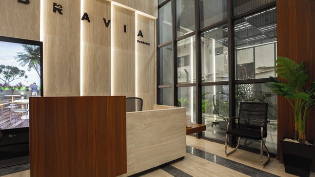 A reception desk with a wooden finish and a glass partition. The reception area has a modern design with a large window providing a view of the outdoors. - Sky Suites BRAVIA RESIDENCE OSU (Accra)