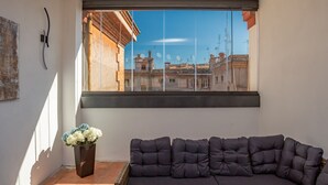 Ein modernes Wohnzimmer mit einem dunkelgrauen Ecksofa, einem kleinen Couchtisch und einer Topfpflanze. Ein großes Fenster bietet Blick auf Gebäude und einen klaren Himmel.