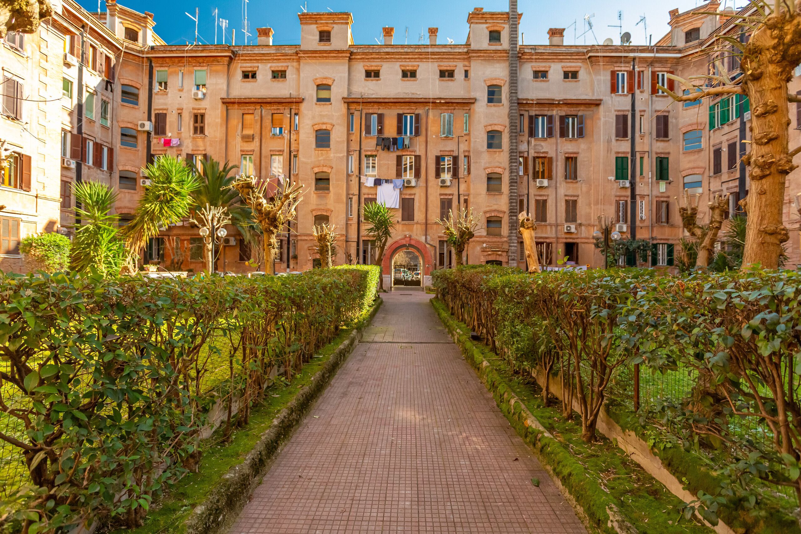 A pathway leading to a building with multiple floors, surrounded by well-maintained hedges and trees.