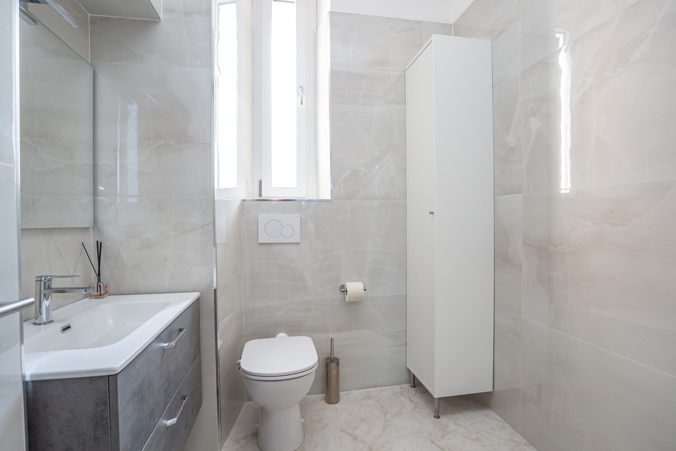 Une salle de bain moderne avec un lavabo, des toilettes et un meuble de rangement blancs, ainsi qu’une fenêtre qui laisse entrer la lumière naturelle.