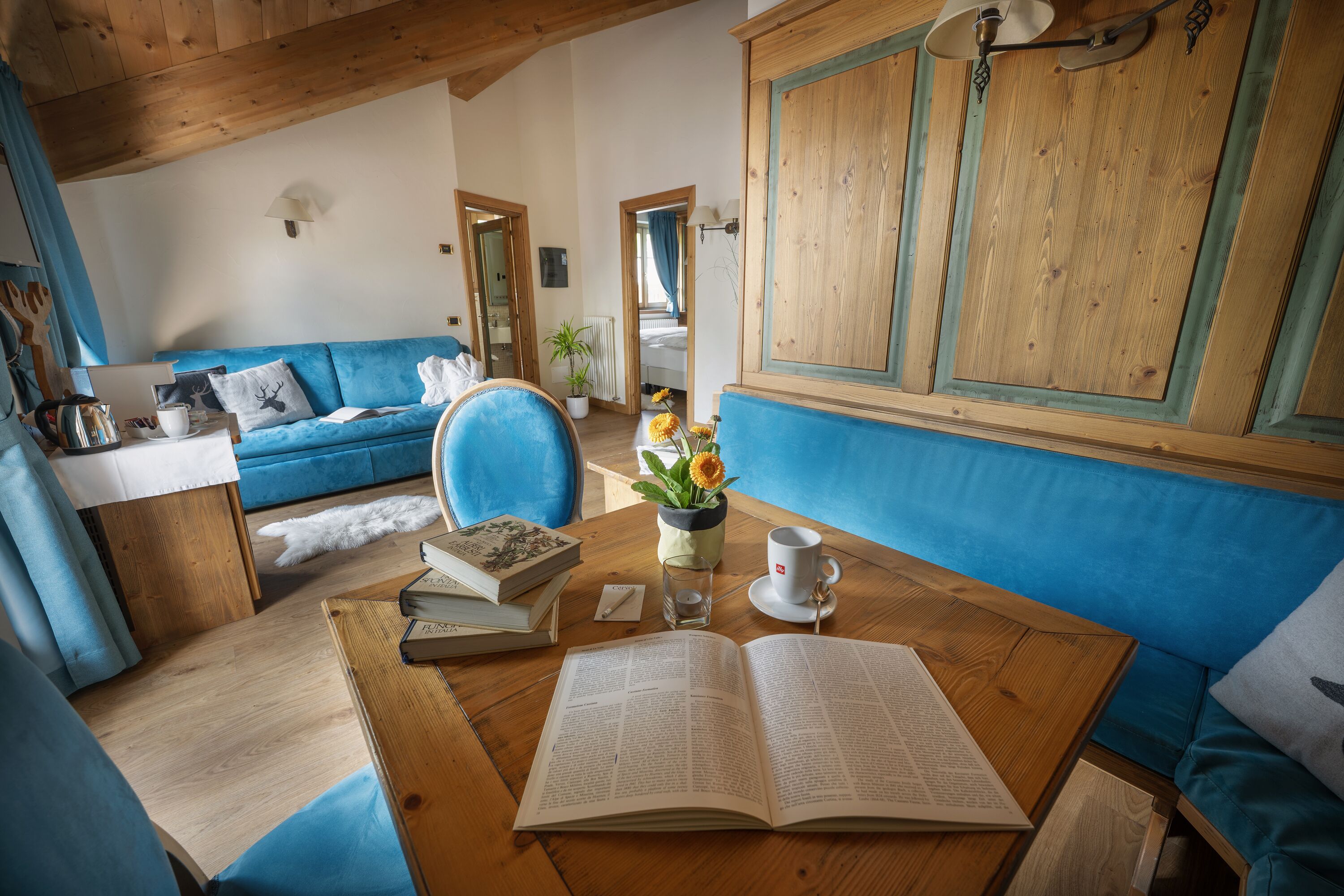 A cozy room with a wooden ceiling, a blue sofa, a wooden table with books and a cup, and a view of a bedroom through an open door.