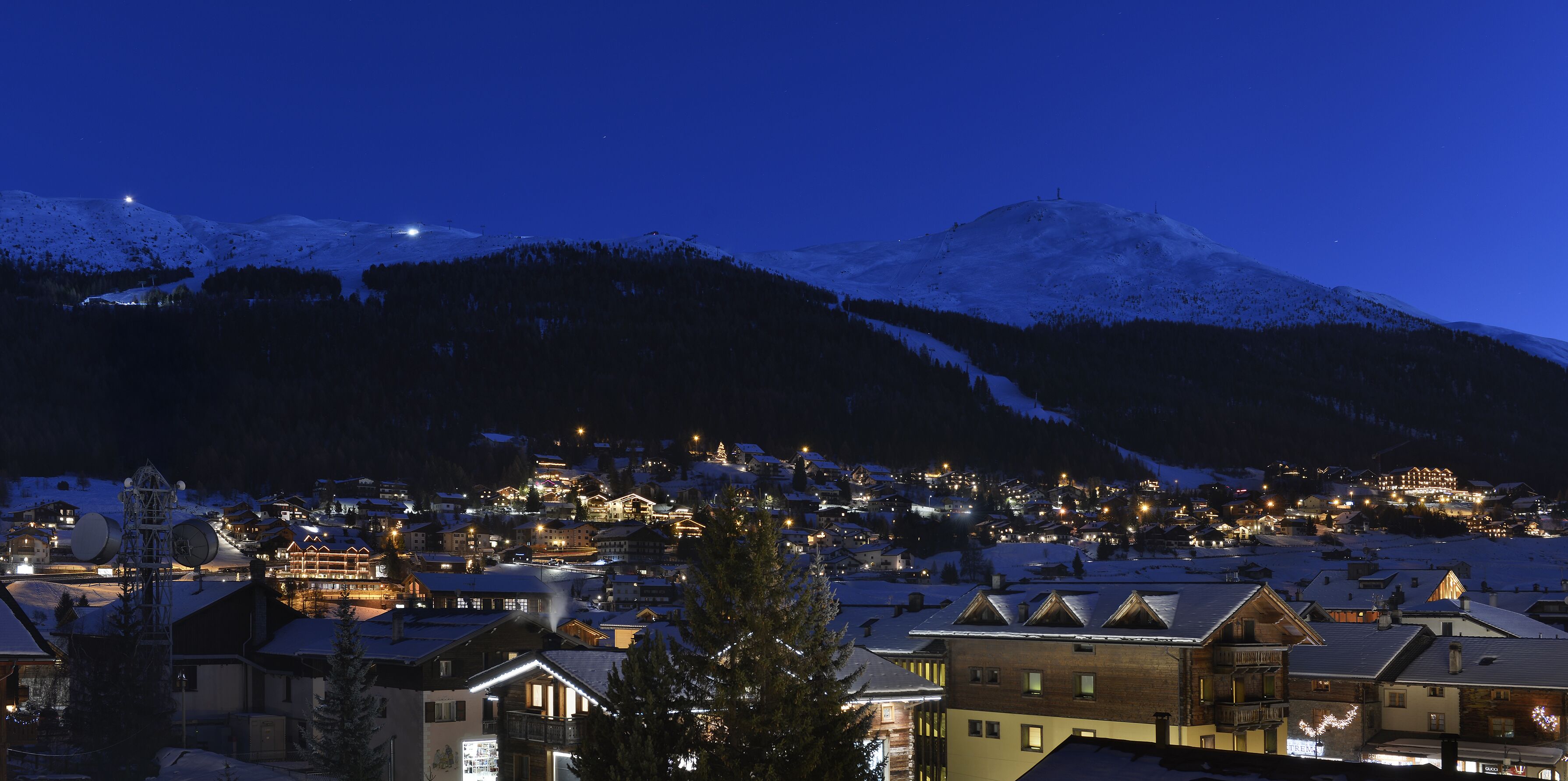 Un pueblo cubierto de nieve al caer la noche, con edificios iluminados y un importante complejo de esquí al fondo.