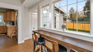 Eine Küche mit Holzausstattung, ein Essbereich mit Tisch und Stühlen und große Fenster mit Blick auf einen Garten mit Holzlattenzaun und Bäumen.