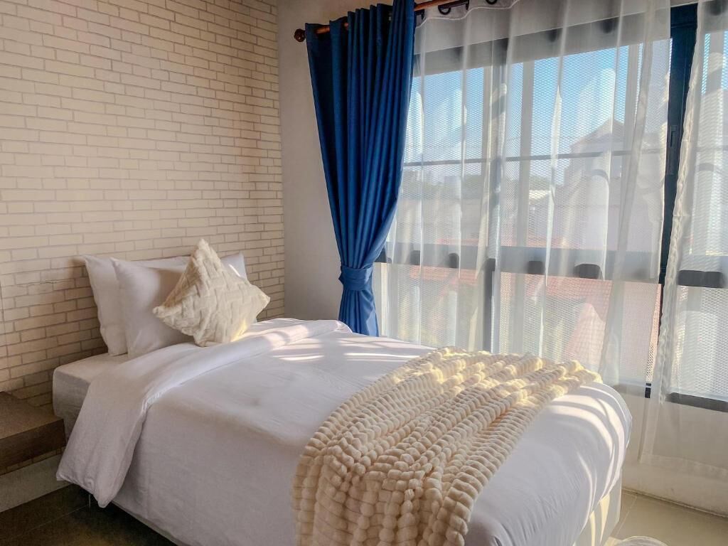 A bedroom with a white bed, a white blanket, and a view of a balcony through sheer curtains.