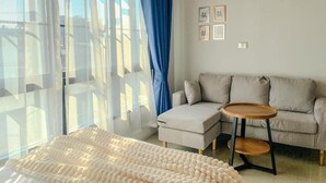 Un dormitorio moderno con una cama, un sofá, una mesita y una ventana con cortinas translúcidas.