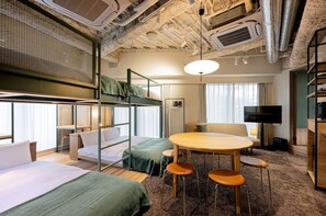 Et hotelværelse med køjesenge, et rundt bord med stole, et fjernsyn og et stort vindue.