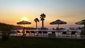 Zona junto a la piscina con sillones reclinables y sombrillas, con vista a un cuerpo de agua y palmeras al atardecer.