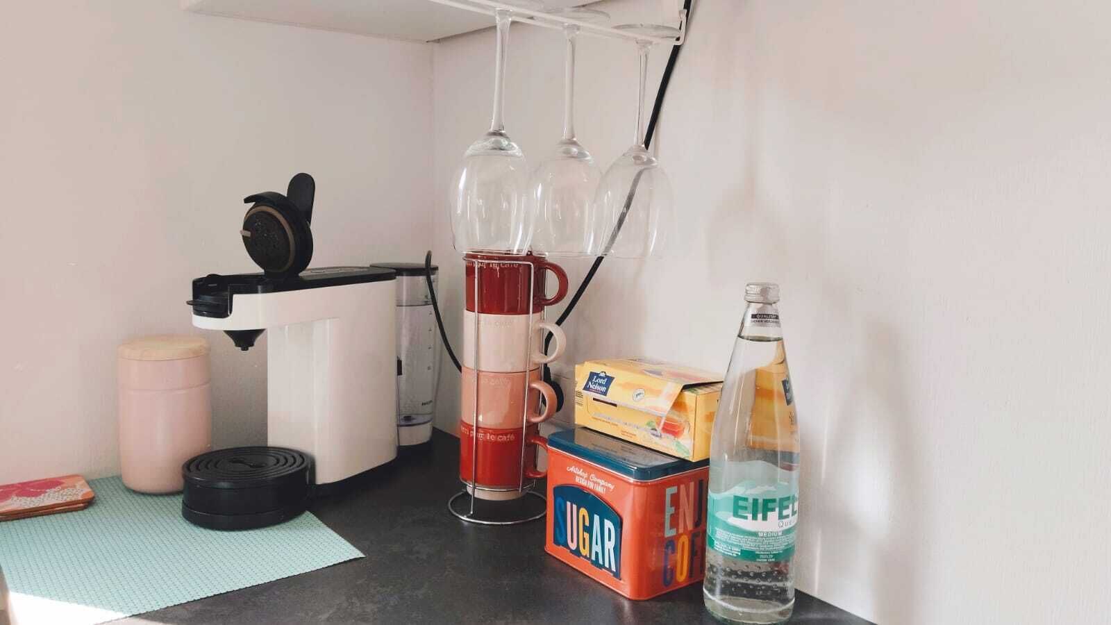 A kitchen counter with a coffee maker, sugar, a box of sugar, a bottle of water, and a pair of headphones.