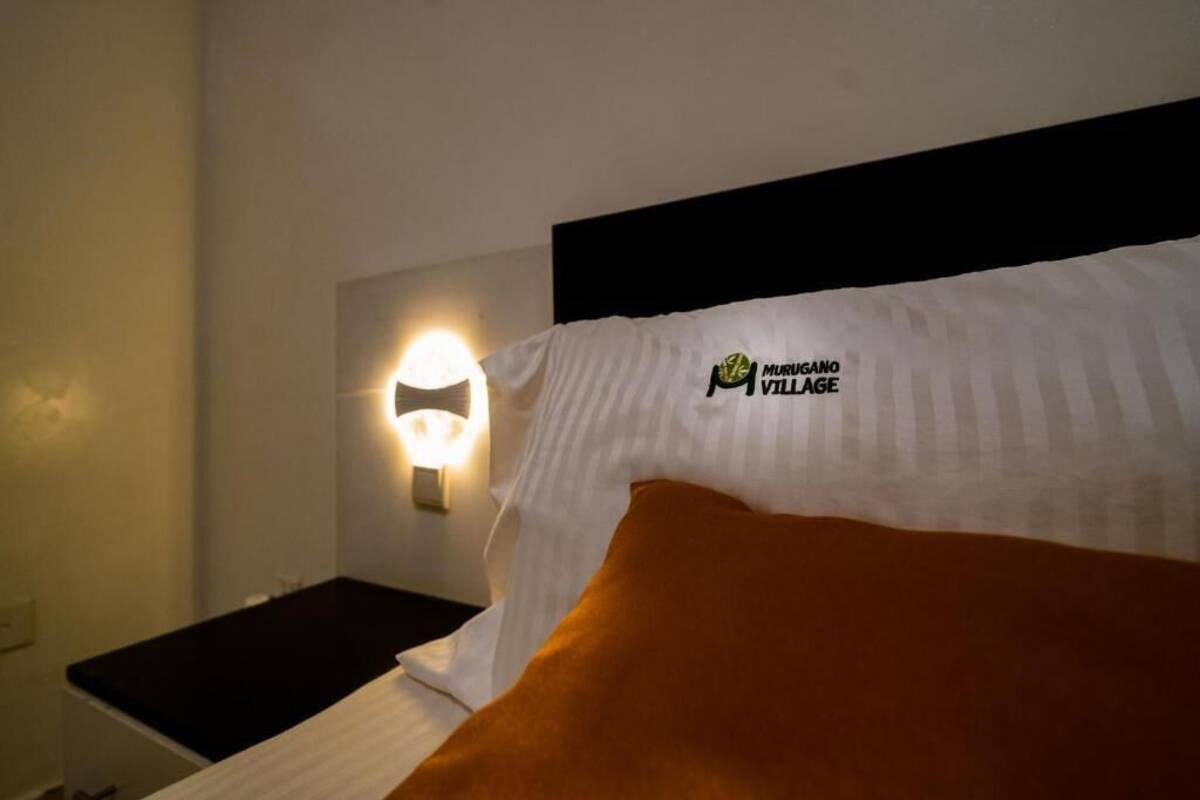 A hotel room with a bed featuring a Murugano Village logo pillow, a bedside lamp, and a black nightstand.