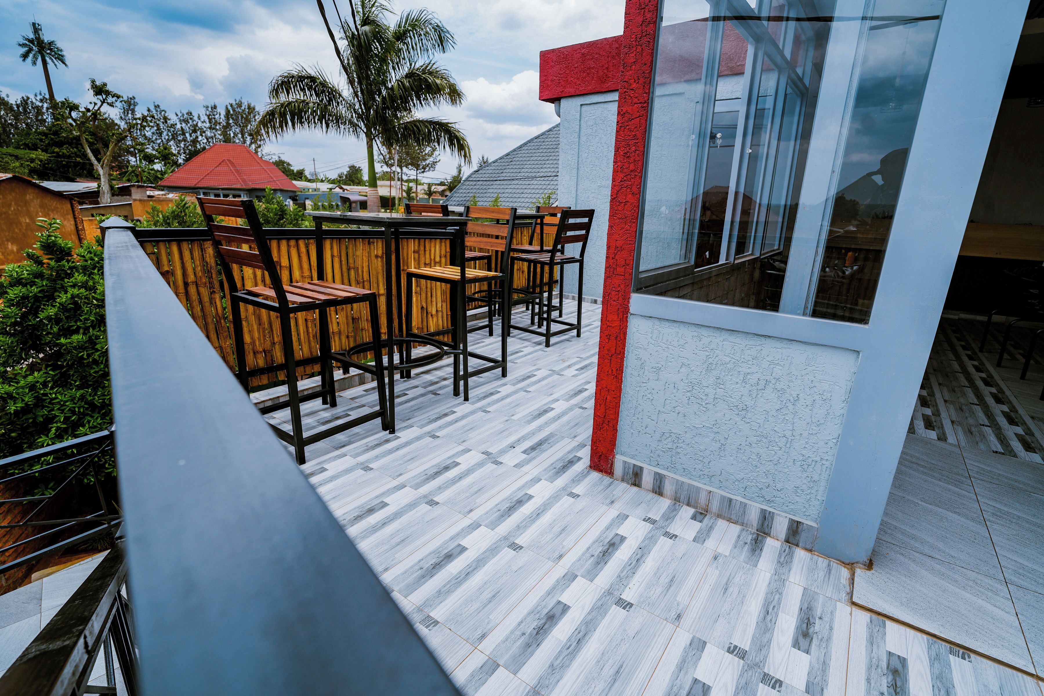 A balcony with wooden chairs and a table, a red accent wall, and a view of the sky and palm trees.