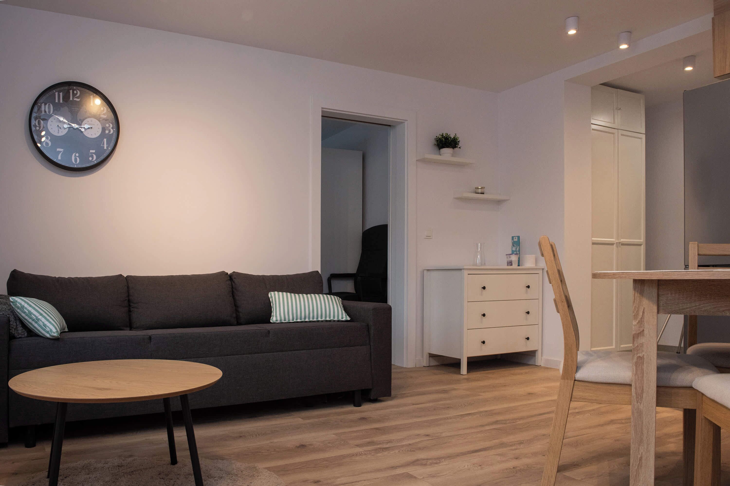 Un salon moderne comprenant un canapé, une table ronde en bois, une horloge, un bureau avec des tiroirs et une chaise.