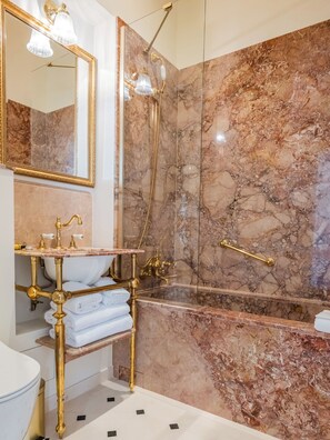 Un baño con pared de mármol, un espejo con marco dorado, un lavabo blanco con detalles dorados y una zona de ducha con mampara de cristal.