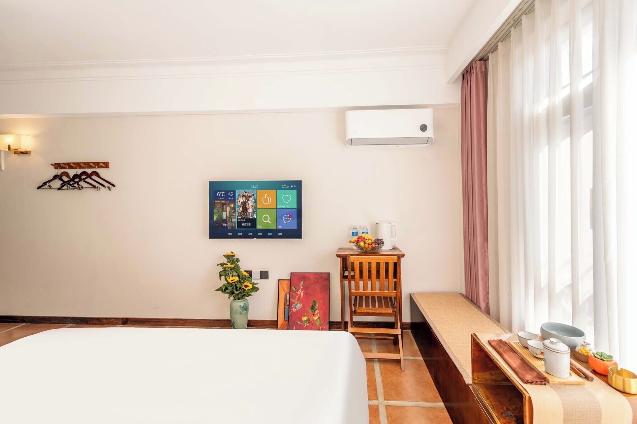 A hotel room with a bed, a wall-mounted TV, a small table with a bowl of fruit, a vase with flowers, and a window with curtains.