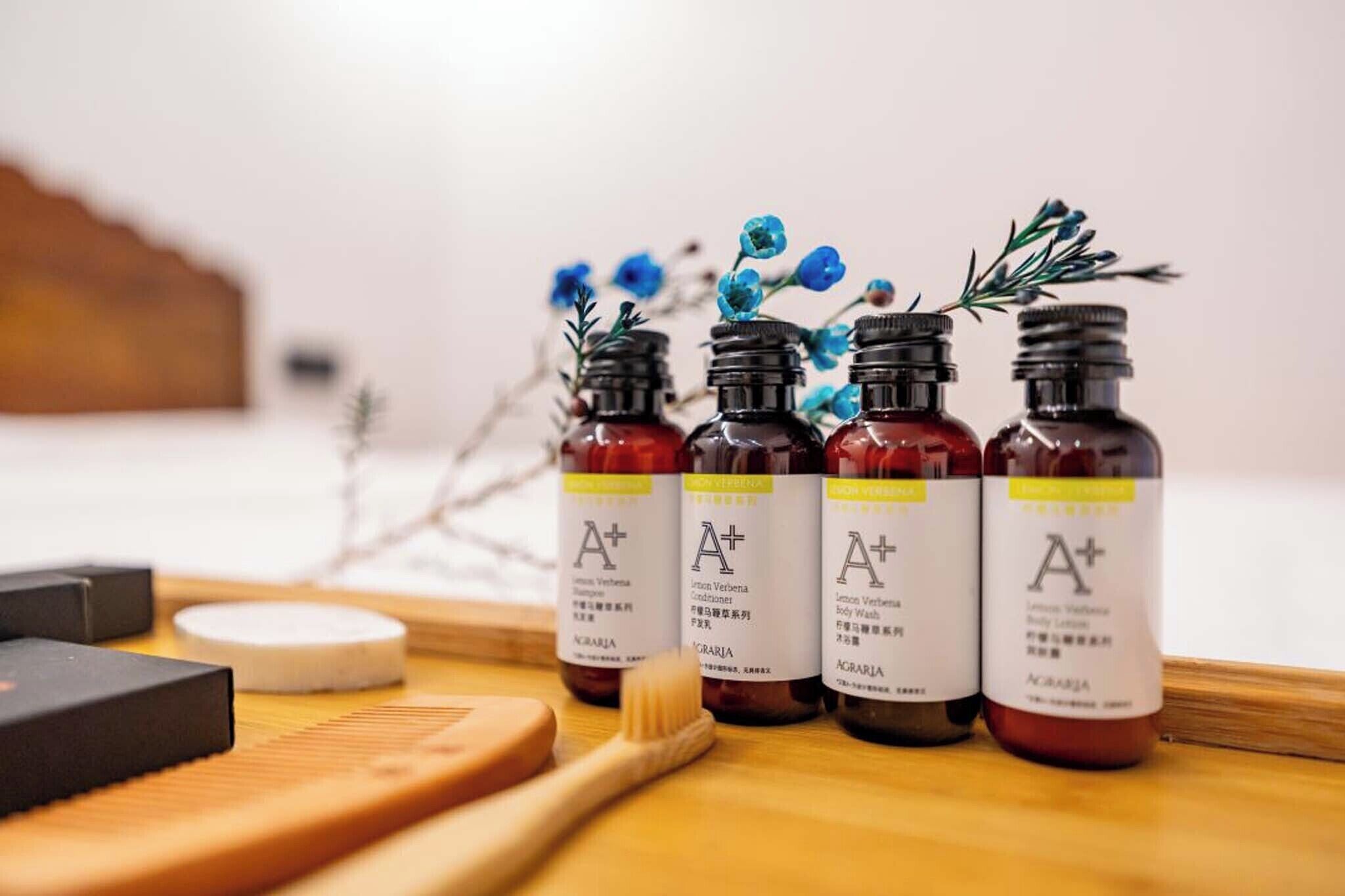 Four bottles of body wash with 'A+' labels, a wooden cutting board, a wooden brush, and some dried flowers.