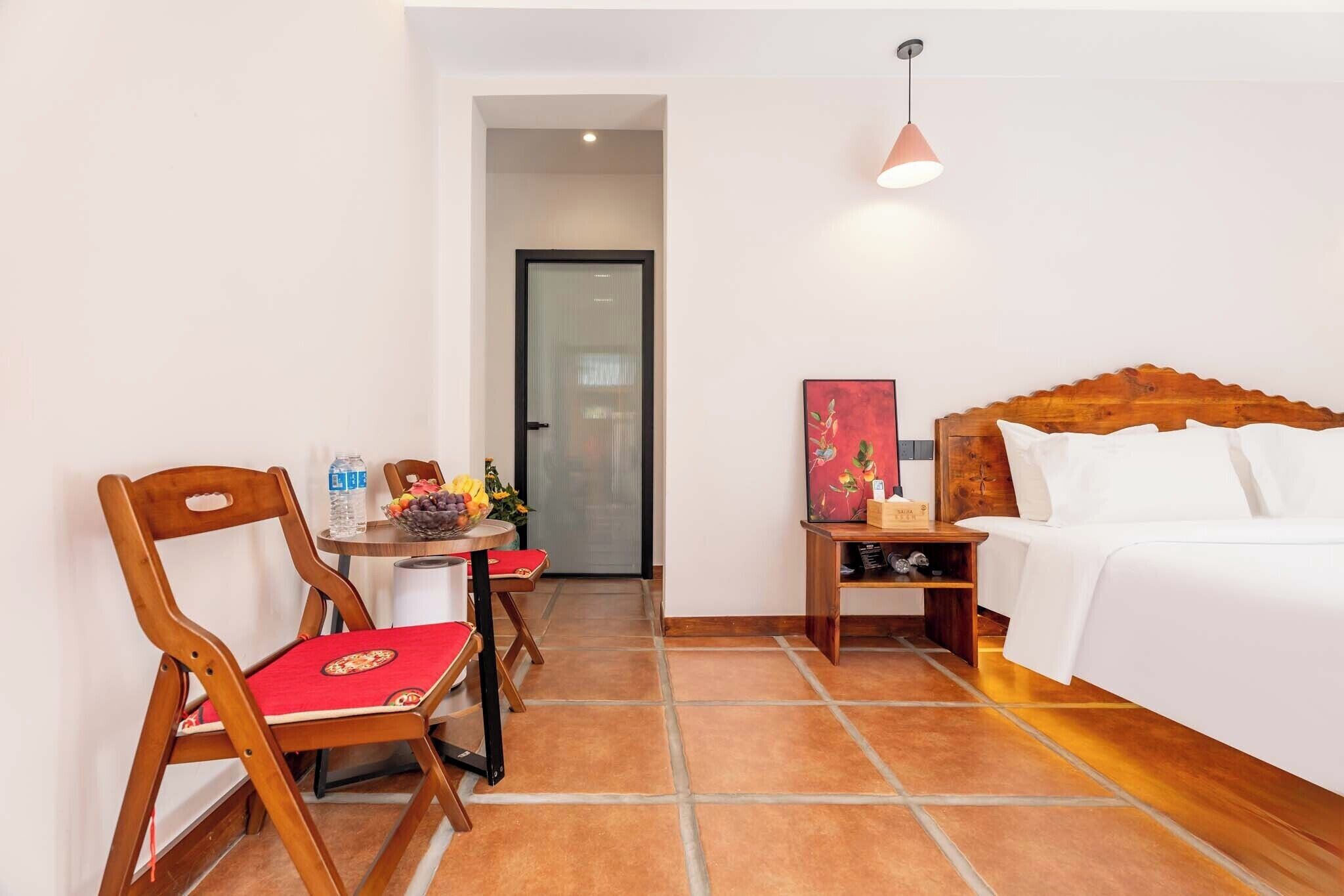 Habitación de hotel con cama, cabecera de madera, una mesita con un florero y dos sillas de madera con cojines rojos.