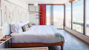 A modern bedroom with a large bed, a view of the city, and a macrame wall hanging. - Native Residence Casa 4 (Playas de Rosarito)